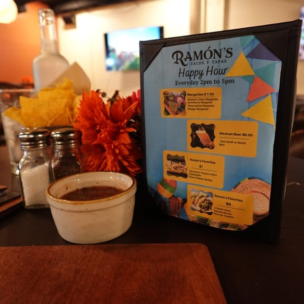 A Ramon's Tacos y Tapas happy hour menu, salsa, tortilla chips, and salt and pepper shakers on a table.